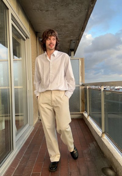 Una persona está de pie en un balcón, vistiendo una camisa blanca de botones, pantalones beige y zapatos negros, con un fondo de pared texturada de color claro.