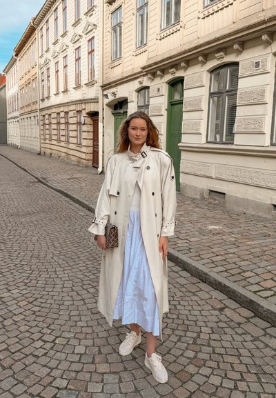 Joven mujer con abrigo blanco tipo trench, vestido azul claro y zapatillas blancas, de pie en una calle empedrada con edificios históricos al fondo.