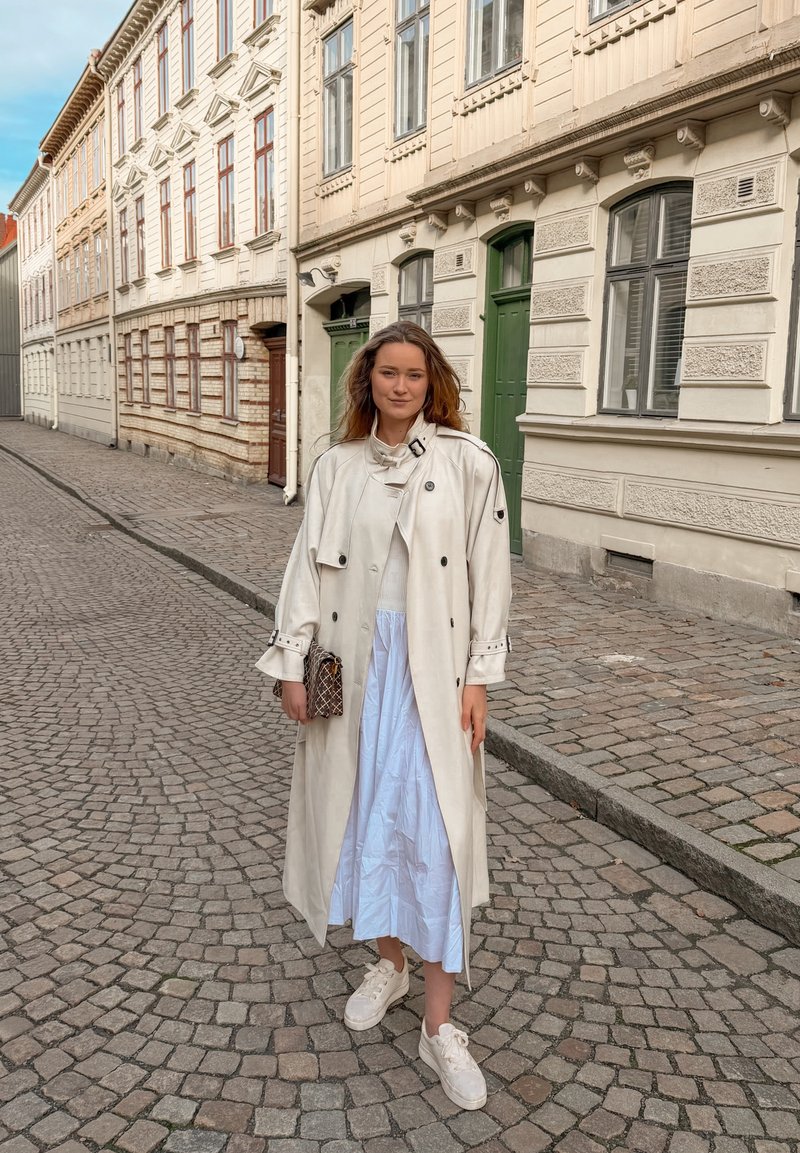 Jeune femme en trench-coat blanc, robe bleu clair et baskets blanches se tient dans une rue pavée avec des bâtiments historiques en arrière-plan.