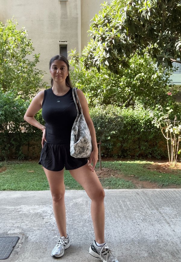 Joven mujer con ropa deportiva negra y zapatillas se encuentra al aire libre sobre hormigón cerca de arbustos verdes, sosteniendo un bolso de hombro con estampado.