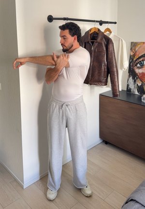 Hombre con camisa blanca y pantalones deportivos grises estira su brazo en una habitación con una chaqueta de cuero y una camisa colgadas detrás de él.