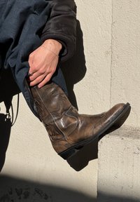 Brūni ādas kovboju zābaki ar dekoratīvo izšuvumu, norādīts purngals un zems kaudzes papēdis, balstīti pret gaišas krāsas sienu.