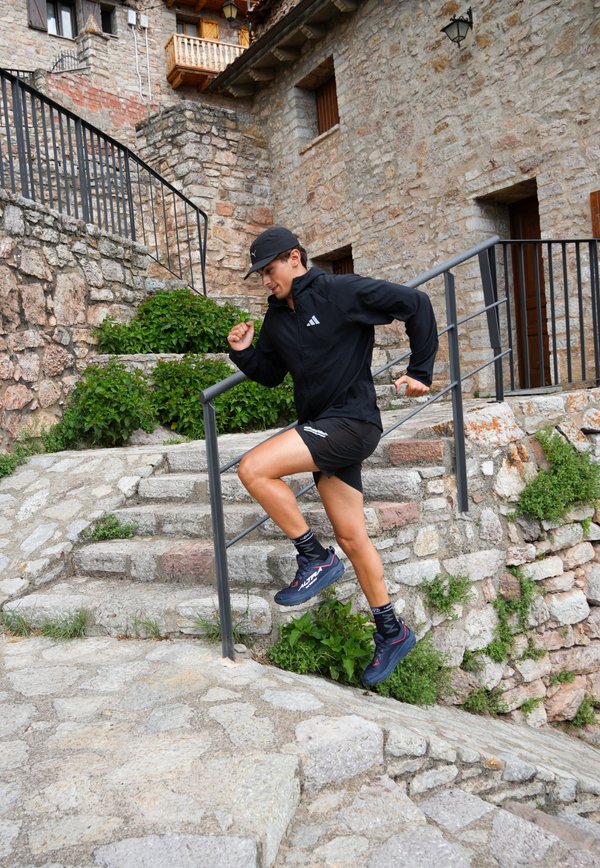 Giovane uomo in abbigliamento sportivo nero che corre su scale di pietra all'aperto vicino a un edificio rustico in pietra con ringhiera in metallo e vegetazione.