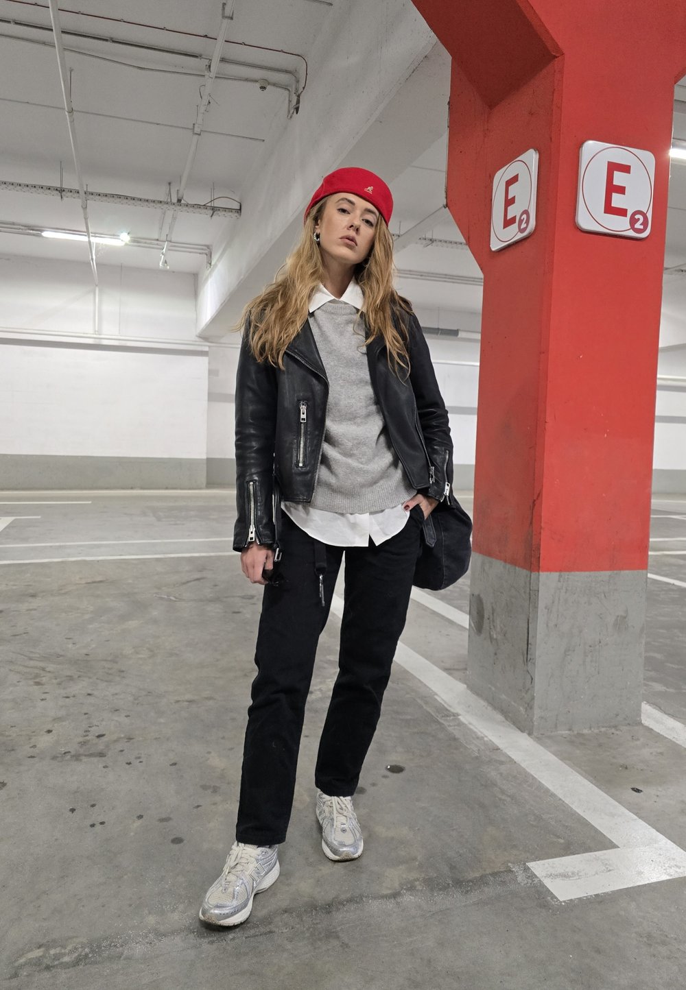Δερμάτινο μπουφάν μηχανής πάνω από γκρι πουλόβερ, λευκό πουκάμισο, μαύρο παντελόνι και ασημένια sneakers. Το κόκκινο καπέλο και η μαύρη τσάντα προσθέτουν αντίθεση.