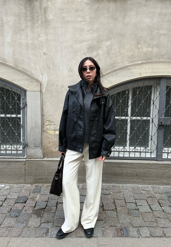 Žena stojaca na kamennej ulici, má na sebe čiernu koženú bundu, sivý sveter, biele nohavice, čierne topánky, slnečné okuliare a drží čiernu kabelku.