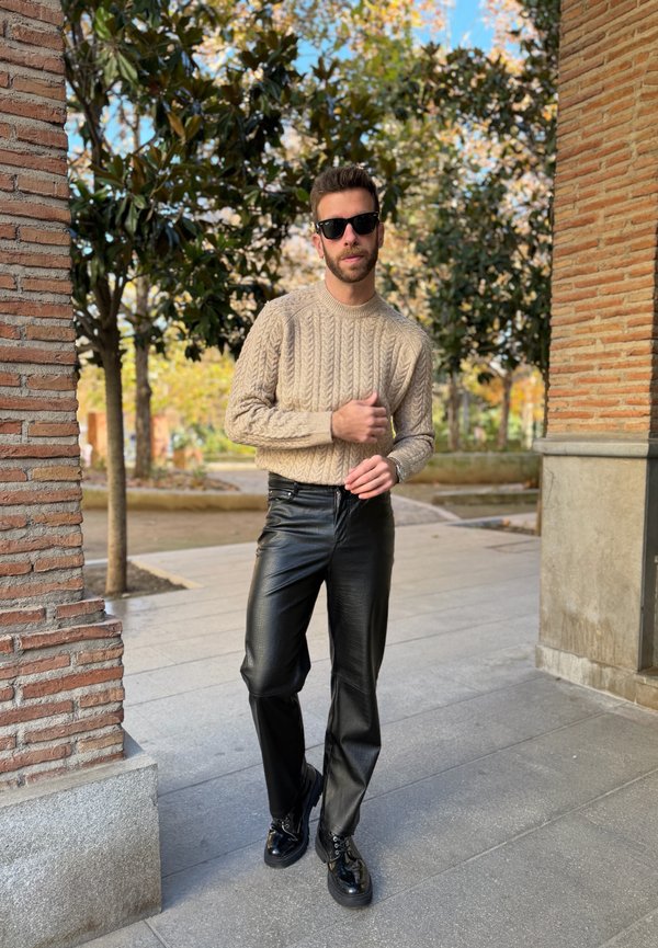 Homme portant des lunettes de soleil, un pull en laine torsadée beige et un pantalon en cuir noir, debout à l'extérieur près de colonnes en briques et d'arbres.