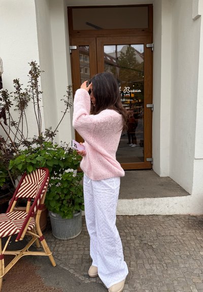 Suéter de punto rosa claro con un corte holgado, combinado con pantalones anchos blancos estampados. El fondo presenta una puerta y plantas en macetas.