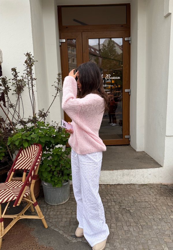 Light pink knitted sweater with a relaxed fit, paired with white patterned wide-leg trousers. The background includes a door and potted plants.
