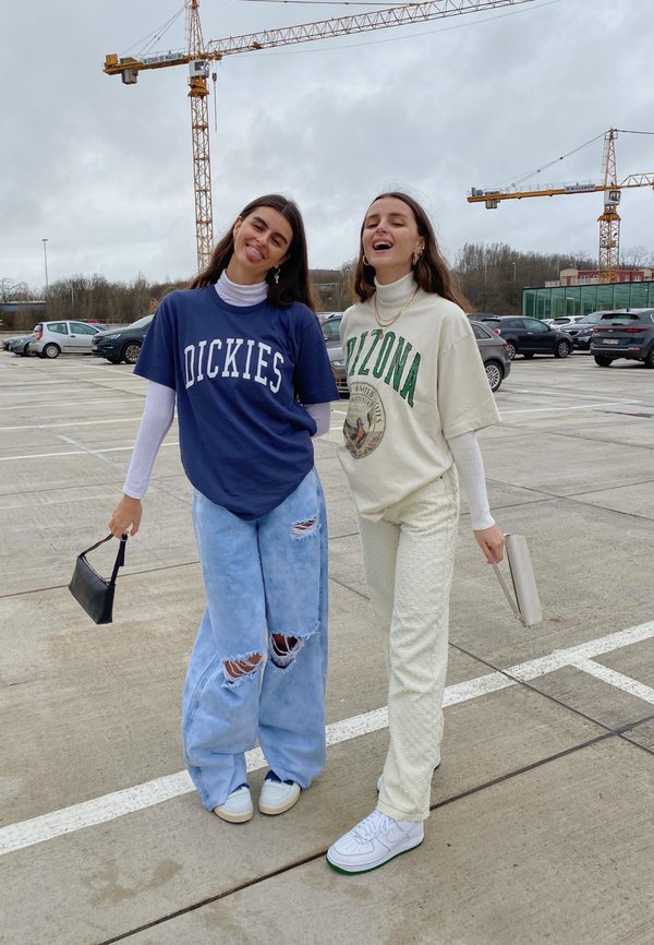 Dos personas visten atuendos informales: una lleva una camiseta azul marino con el texto "Dickies", jeans azul desgastados y zapatillas blancas; la otra viste una camiseta beige de "Arizona" con pantalones claros y texturizados y una bolsa pequeña.