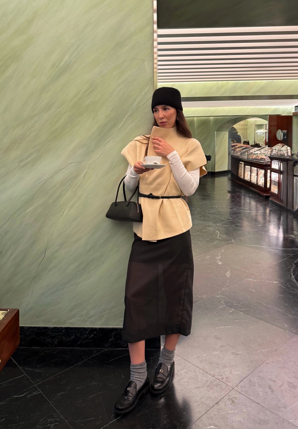Beige wool wrap top paired with a black midi skirt, black loafers, and grey socks. Set against a green marble background.