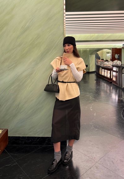 Top beige de lana tipo envolvente combinado con una falda midi negra, mocasines negros y calcetines grises. Fondo de mármol verde.