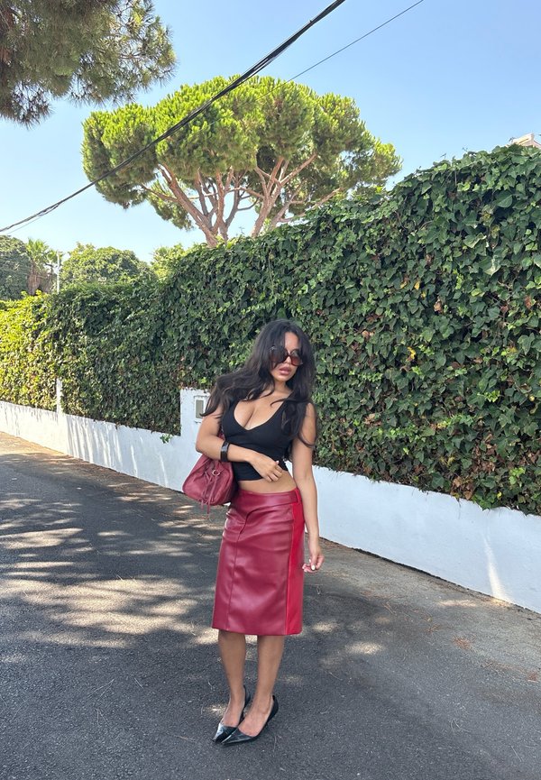Femme portant un top noir, une jupe en cuir rouge et des lunettes de soleil, debout dans la rue à côté d’un haut mur vert couvert de lierre en plein jour.