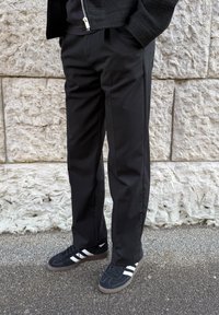 Personne portant un pantalon noir, une veste noire avec une fermeture éclair et des baskets noires avec des bandes blanches, debout sur un trottoir contre un mur en pierre texturé.