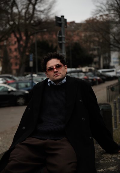 Hombre con abrigo oscuro, suéter y gafas de sol rojas apoyado en un borde de piedra, con coches estacionados y árboles en un entorno urbano nublado.