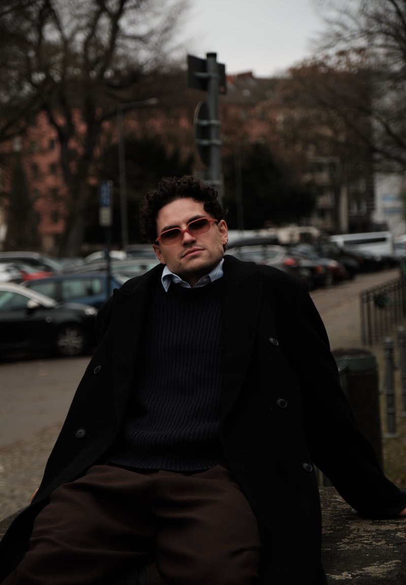 Homme portant un manteau sombre, un pull et des lunettes de soleil rouges, appuyé sur un rebord en pierre, avec des voitures garées et des arbres dans un cadre urbain nuageux.