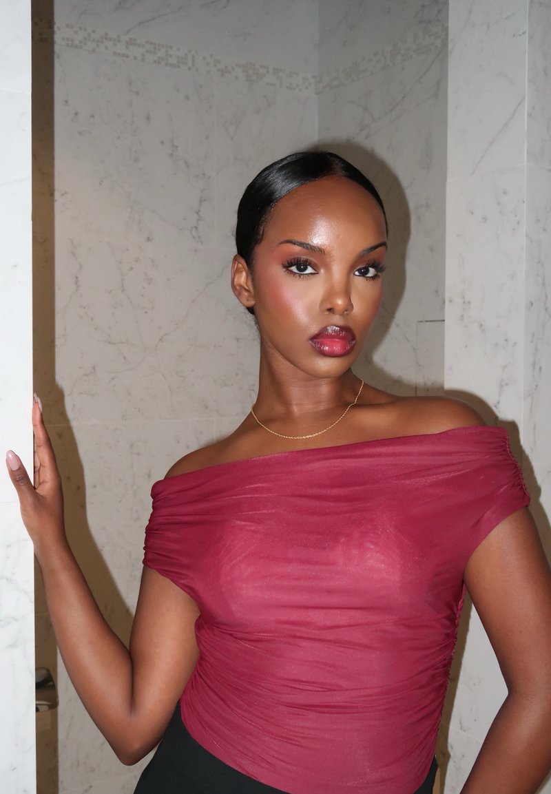 Femme aux cheveux lisses portant un haut bordeaux transparent à épaules dénudées et un collier en or, debout devant un mur en marbre blanc.