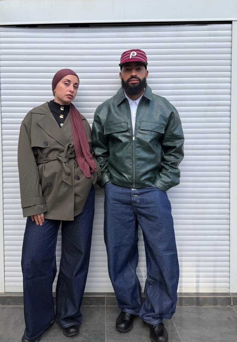 Deux personnes se tiennent devant un volet blanc ; l'une porte un trench-coat vert et un foulard mauve, l'autre une veste en cuir verte et une casquette bordeaux.