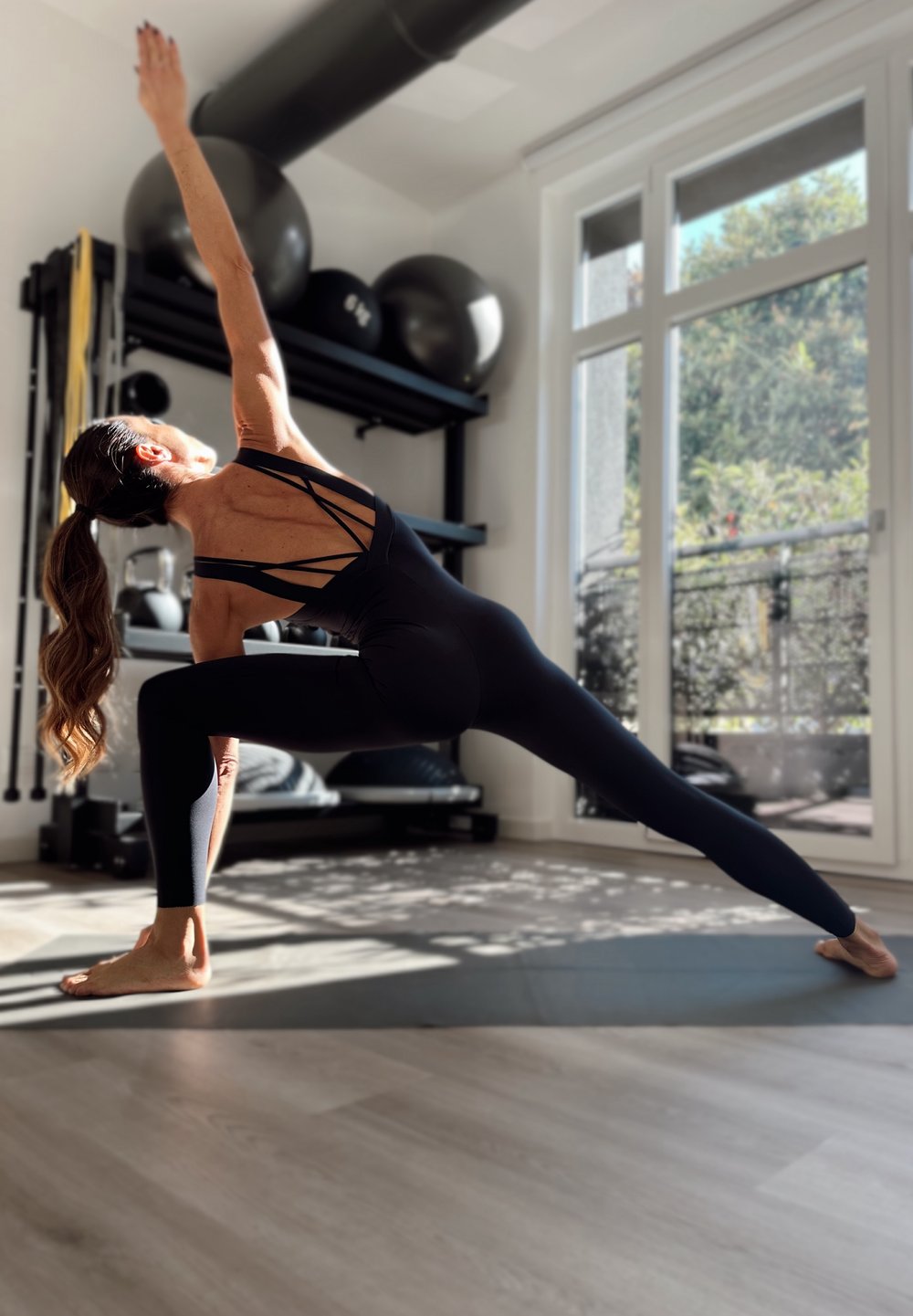 Ženska v črni telovadni obleki se razteza v jogijski pozi na podlogi v svetli sobi z vadbenimi žogami in velikimi okni.