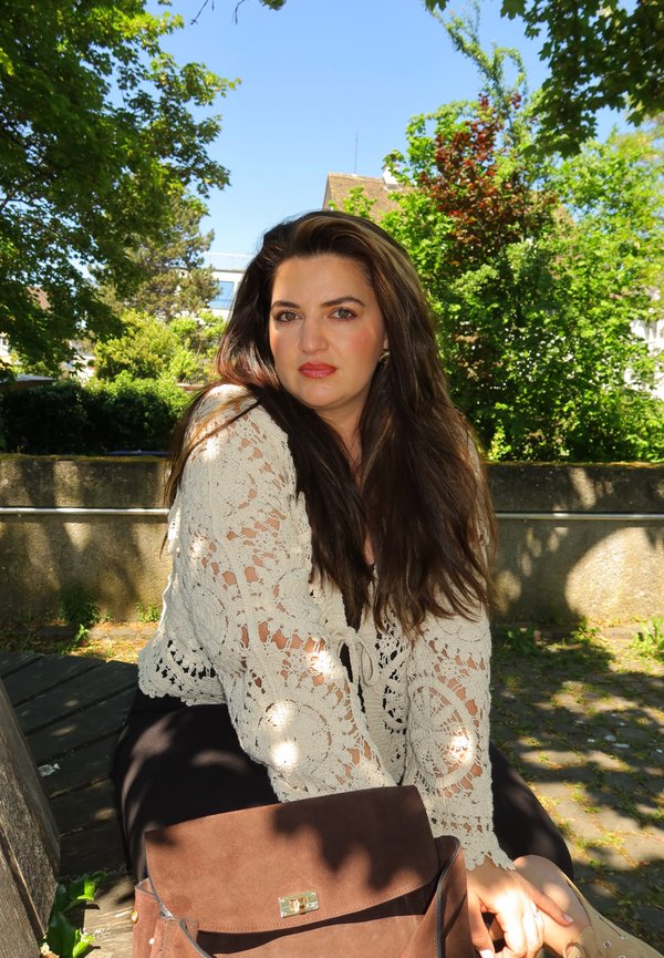 Femme aux longs cheveux bruns portant un haut en dentelle blanc, assise sur un banc en bois en plein air avec des arbres verts et un sac marron devant.
