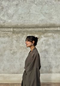 Vrouw met donker haar in een knot, grote zilveren oorbellen en een losse bruine jas, staand zijwaarts tegen een gestructureerde betonnen muur.