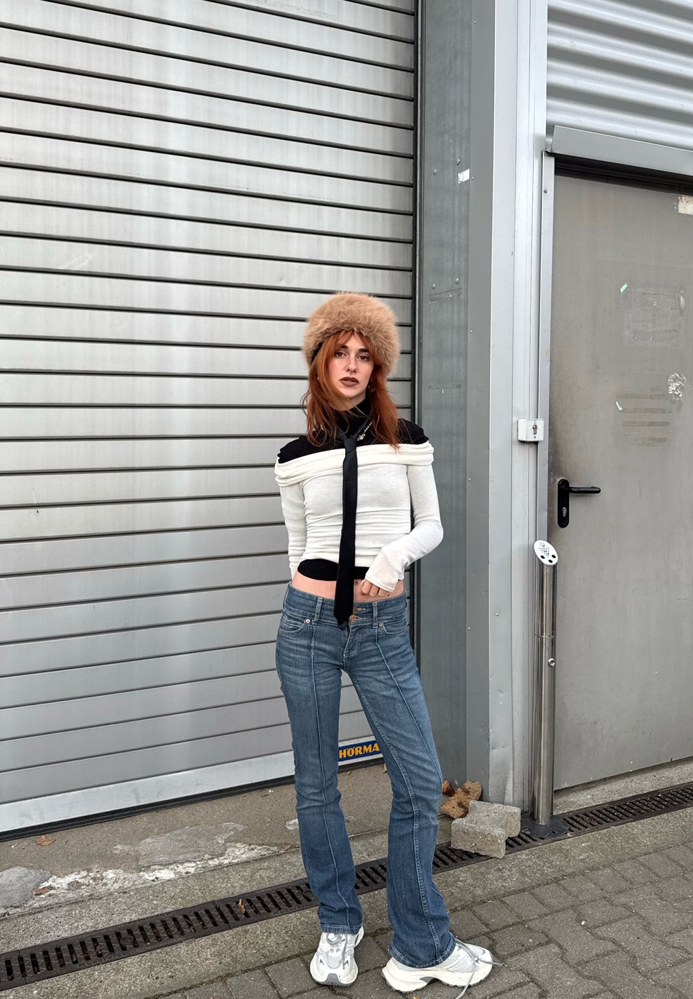 Bluză albă cu mâneci lungi și accente negre, cravată neagră, blugi evazați albaștri și pălărie bej din blană. Fundalul prezintă un panou vertical gri.