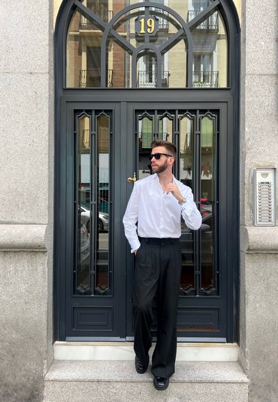 Camisa blanca de botones, pantalones negros con pliegues y zapatos negros. De pie frente a una puerta negra decorativa con parte superior en arco y el número 19.
