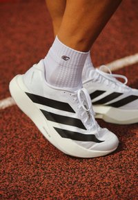 Hvite atletisk sko med tre svarte striper, strukturert overdel og hvite snørebånd. Brukt med en hvit sokk, satt mot en rød løpebane.