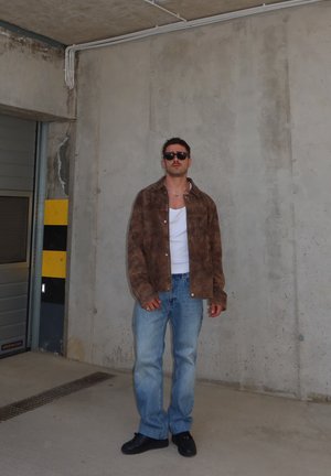 Hombre con gafas de sol, chaqueta marrón, camisa blanca, vaqueros azules y zapatos negros, apoyado contra una pared de hormigón en un aparcamiento cubierto.