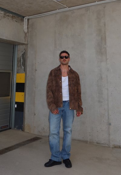 Hombre con gafas de sol, chaqueta marrón, camisa blanca, vaqueros azules y zapatos negros, apoyado contra una pared de hormigón en un aparcamiento cubierto.