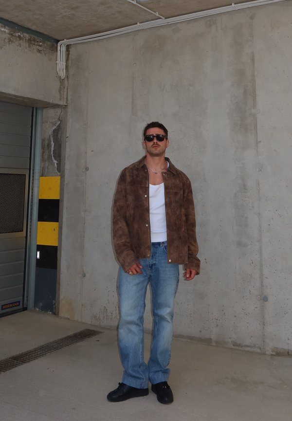 Άνδρας με γυαλιά ηλίου, καφέ μπουφάν, λευκό πουκάμισο, μπλε τζιν και μαύρα παπούτσια, στέκεται δίπλα σε τσιμεντένιο τοίχο σε κλειστό χώρο στάθμευσης.