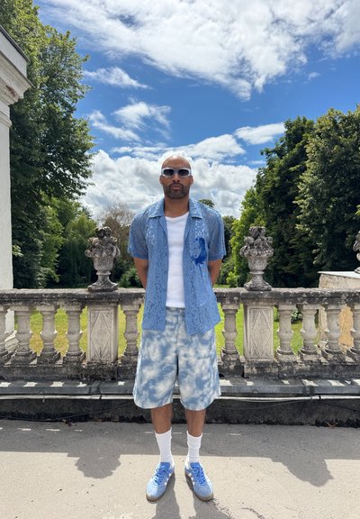 Camisa de encaje azul claro de manga corta sobre una camiseta blanca, combinada con pantalones cortos estampados de nubes. Calcetines blancos y zapatillas azul claro completan el look.