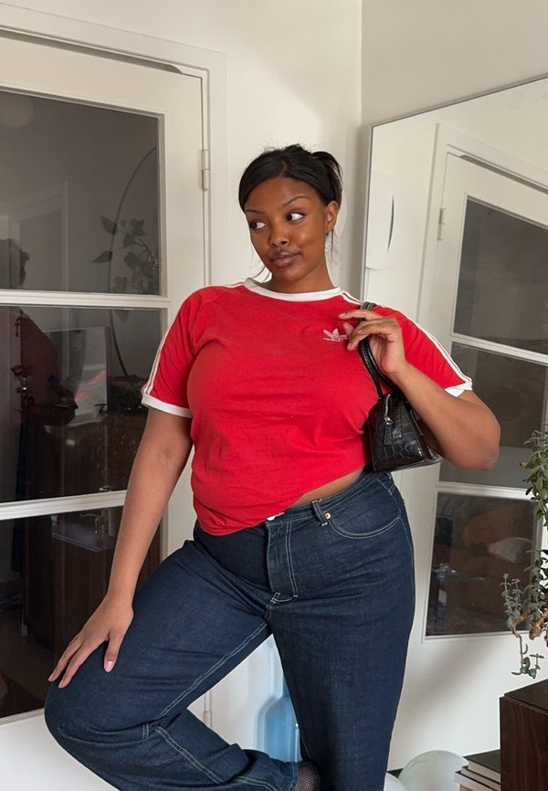 Kvinde iført en rød Adidas t-shirt og mørke jeans poserer indendørs med en sort håndtaske over skulderen.