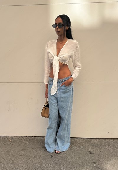 Blusa blanca con cuello en V y lazada en el frente, de mangas largas, combinada con jeans de mezclilla sueltos de color azul claro. Un bolso negro y unas gafas de sol completan el look.