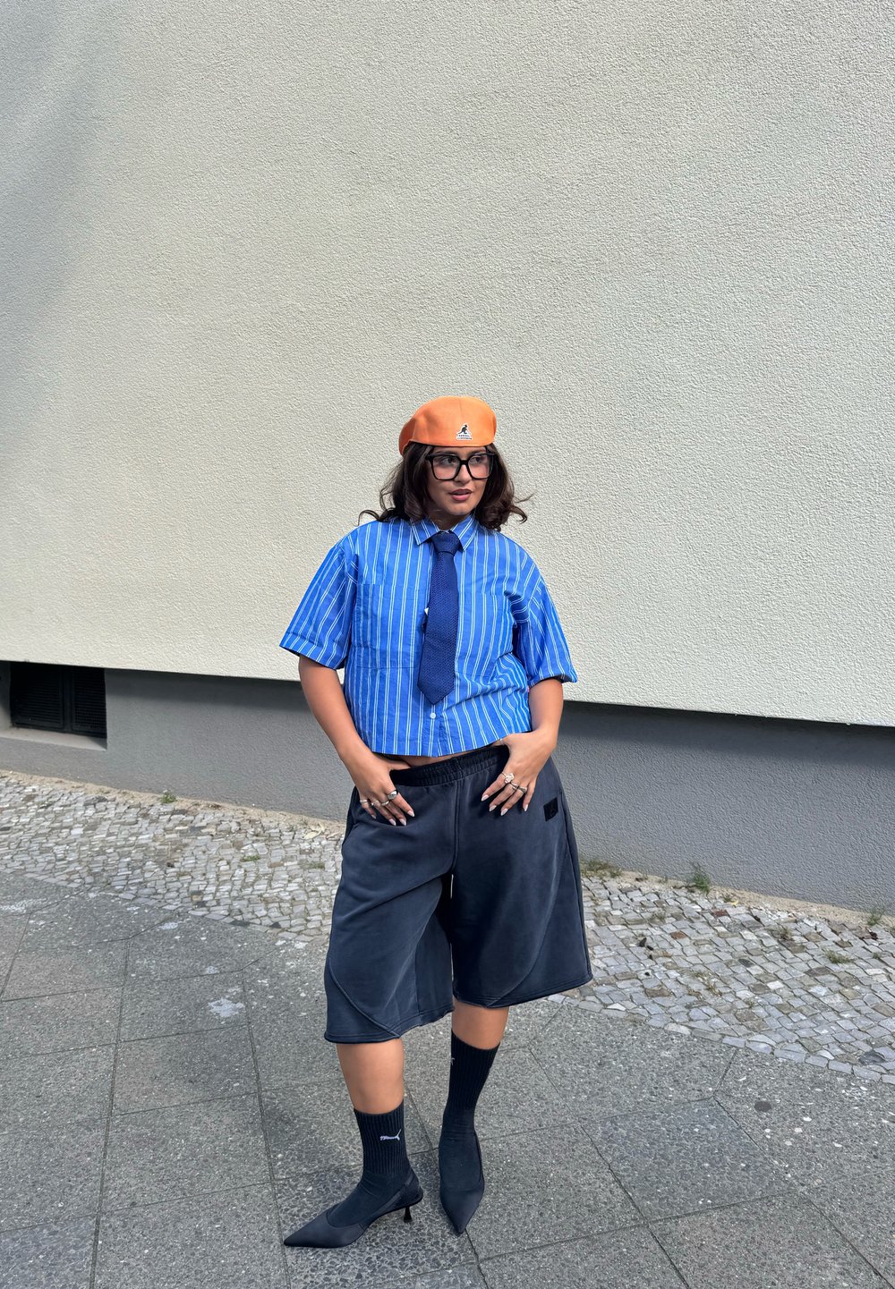 Zilā strīpainā īsā piedurkņu krekls, tumši šorti, melnas augstpapēžu kurpes, potītes zeķes un oranžs berets ar logotipu.