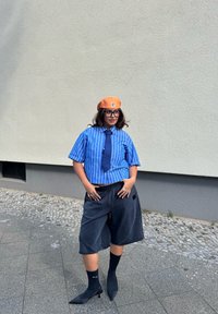 Zilā strīpainā īsā piedurkņu krekls, tumši šorti, melnas augstpapēžu kurpes, potītes zeķes un oranžs berets ar logotipu.