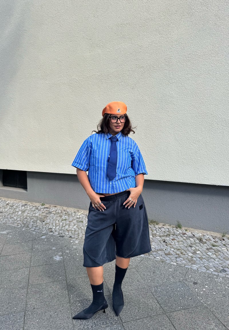 Camicia a righe blu a maniche corte, pantaloni corti scuri, scarpe con tacco nere, calzini alla caviglia e un berretto arancione con un logo.