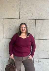 Vínový rebrovaný sveter so širokým výstrihom a textúrovaným vzorom; v kombinácii s hnedými manžestráckymi nohavicami a hnedou kabelkou. Pozadie je hladká kamenná stena.