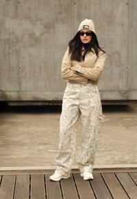 Μπεζ κοντό τζάκετ, ναυτικές μπλούζες, ανοιχτό παντελόνι με σχέδιο φιδιού και φαρδιά πόδια, λευκά αθλητικά παπούτσια και μπεζ σκουφάκι. Τα αξεσουάρ περιλαμβάνουν σκούρα γυαλιά ηλίου.