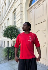 Κόκκινη κοντομάνικη μπλούζα με πολύχρωμο γραφικό και κείμενο στο στήθος. Το υλικό φαίνεται μαλακό με άνετη ε�φαρμογή. Μαύροι φαρδιοί παντελόνες.