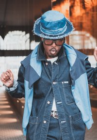 Denim jacket with contrasting blue accents and pocket details, paired with a frayed denim bucket hat and sunglasses, set against an urban background.