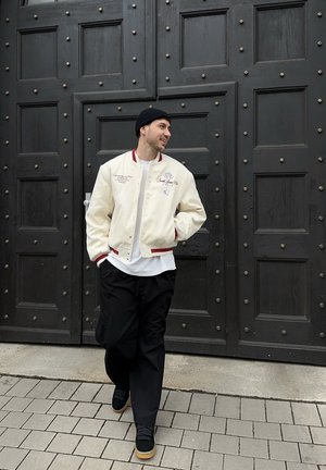 Hombre con chaqueta bomber crema, gorro negro y pantalones negros caminando por una acera de baldosas frente a grandes puertas de madera negras.