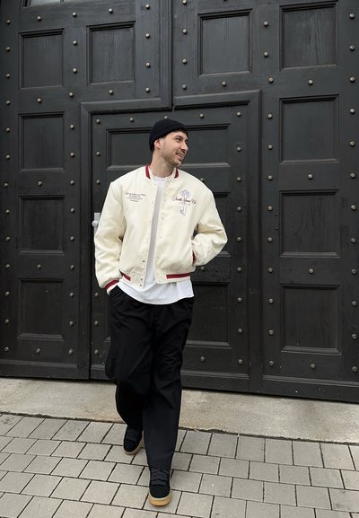 Hombre con chaqueta bomber crema, gorro negro y pantalones negros caminando por una acera de baldosas frente a grandes puertas de madera negras.