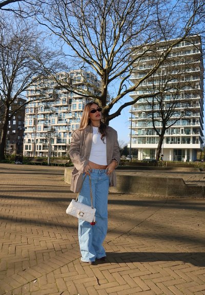 Mujer con gafas de sol, blazer de cuadros, top corto blanco y vaqueros azules, de pie en un área pavimentada sosteniendo un bolso blanco, con árboles sin hojas y edificios altos detrás.