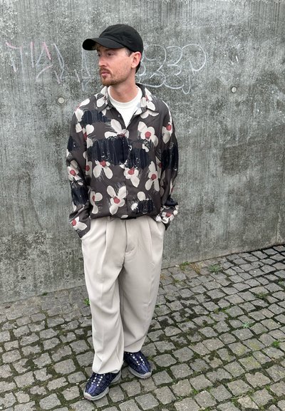 Camisa de manga larga con estampado floral en tonos oscuros, combinada con pantalones plisados beige claros y zapatillas navy, contra un fondo de pared de hormigón.