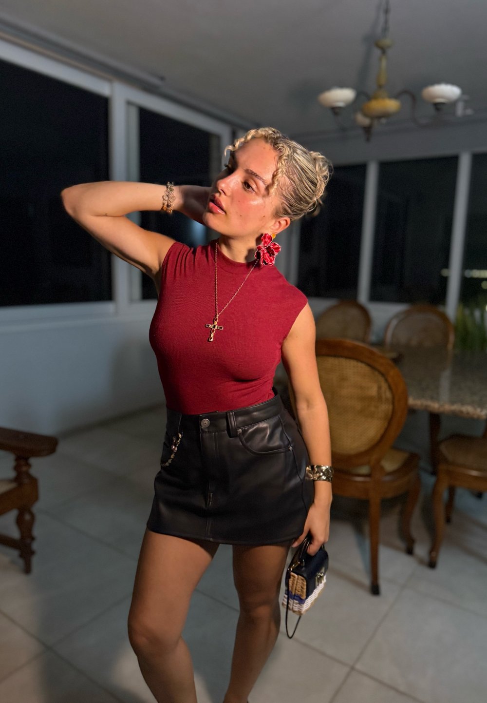 Röd ärmlös topp, svart läder minikjol och mönstrad handväska. Lockigt håraccessoar och guldsmycken. Neutral bakgrund med möbler.