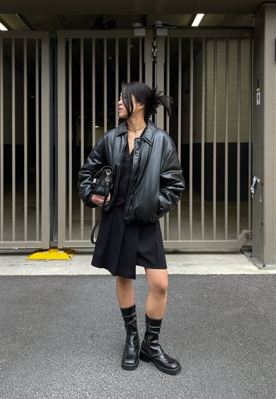 Chaqueta bomber de cuero negro sobre un vestido chemisier negro, combinada con botas hasta la rodilla negras y un bolso pequeño. Fondo neutro.