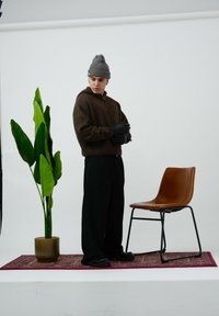 Brązowy sweter zapinany na zamek, czarne rękawiczki, czarne spodnie o szerokich nogawkach oraz czarne buty. Obok roślina i brązowe krzesło na wzorzystym dywanie.