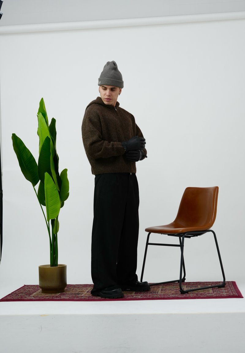 Brauner Strickpullover mit Reißverschluss, schwarze Handschuhe, schwarze Hosen mit weitem Bein und schwarze Schuhe. Neben einer Pflanze und einem braunen Stuhl auf einem gemusterten Teppich.