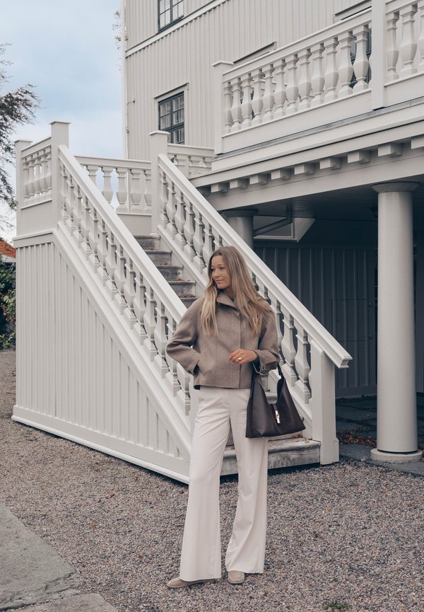 Una mujer con una chaqueta de color topo y pantalones blancos está de pie junto a una escalera de madera blanca y lleva un bolso oscuro.