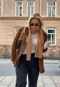 Kobieta w okularach przeciwsłonecznych, brązowej kurtce i beżowym szaliku spaceruje po chodniku w mieście obok beżowego budynku mieszkalnego z białymi oknami.